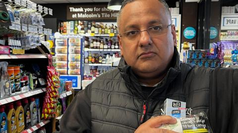 Paul Cheema is a man in his 50s wearing glasses with grey and black hair. He is standing in his petrol station in front of the tills and he is holding illegal tobacco products including a packet of 20 cigarettes and a bag of tobacco.