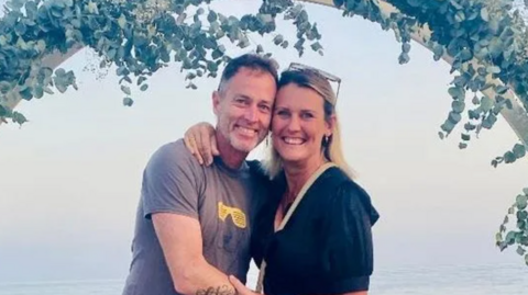 A blonde woman and a grey haired man smile at a camera. They are hugging. They are standing in front of the sea 
