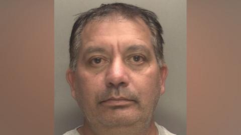 A police mugshot of Harminder Mattu. He is a man with grey hair looking directly at the camera. He is standing against a light-coloured backdrop.