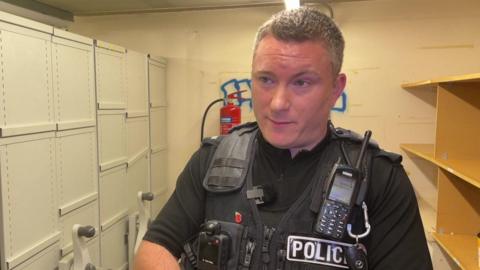 A man wearing black clothing, with a mobile device attached and a badge saying 'POLICE' on his left chest