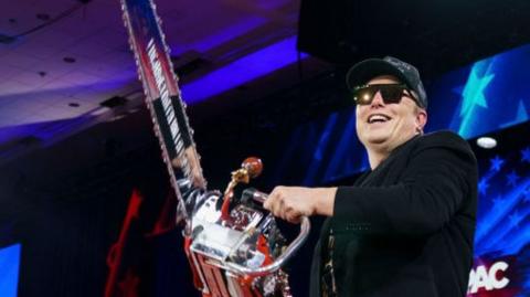 Elon Musk - wearing a hat and sunglasses and grinning broadly - wields a chainsaw on stage at the CPAC conference near Washington this week