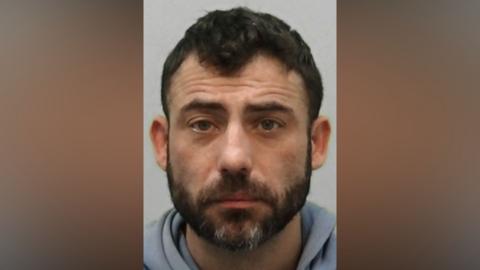 Mugshot of Rayne. He has short black hair and a thick black beard with white patches.