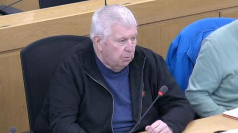 Councillor Mick Glynn, an older man with short, white hair and a black jacket and blue T-shirt, sits at a wooden desk with a microphone.