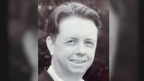 A black and white photograph of Simon Scott wearing a dark suit and a white shirt.