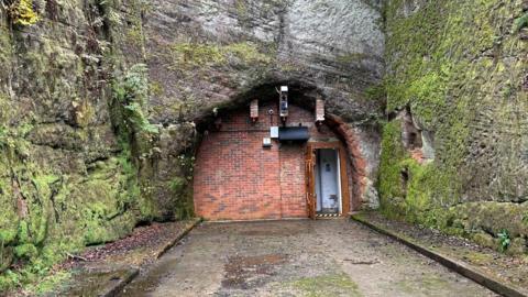 A mossy green rock face with a red brick building built into the side of it. There is a small brown door on the right hand side and path leading up to it.