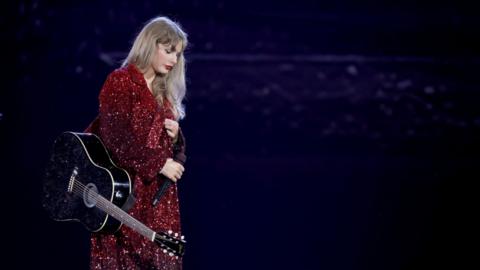 Taylor Swift is wearing a sparkly red dress and stands with a guitar hanging to her right hand side.