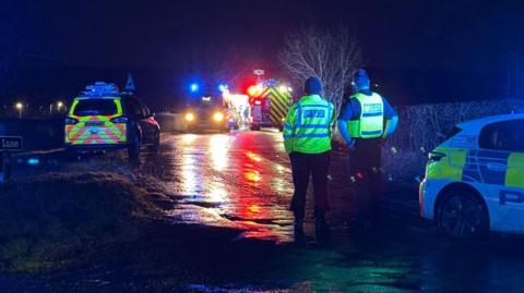 A dark road, with emergency service crews at the scene. Two police officers are stood with their backs to the camera. Headlights are reflecting off a wet road surface. 