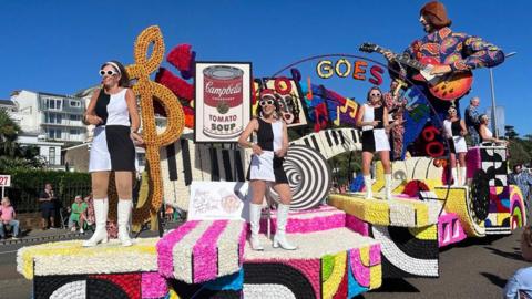 A brightly decorated parade float covered in flowers features oversized piano keys, musical notes, a Campbell’s soup can image, and a large guitar-playing figure. Several performers in matching black-and-white outfits and white boots stand on the float during a sunny outdoor parade.