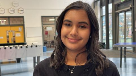 A smiling Sienna Ahir has long dark brown hair and is wearing a black jumper and a necklace with a single pearl. She is standing in the lobby of a building with table football and table tennis table seen in the background. 
