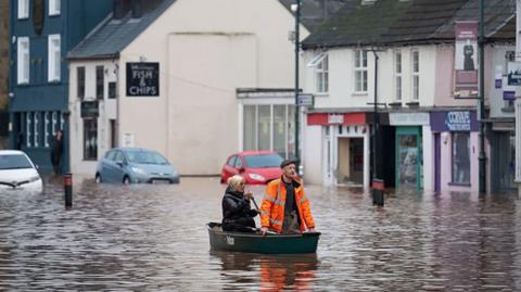 A man and woman pictured using a small boat to navigate the floodwater on what appears to be a high street, The woman is wearing a black jacket and the man is wearing a high-vis jacket