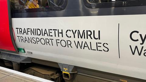 A close-up shot of the side of a train. It is light grey and has the words Trafnidaeth Cymru, Transport for Wales written on it in large black text