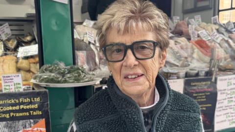 An older woman is looking at the camera, her expression neutral. She is wearing thick black rimmed glasses, and has short blonde hair. She is also wearing a green gilet and a patterned jumper. She's standing in front of her fish stall.