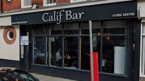 The bar has a black facade with a white sign. It has large square windows and its phone number on the top right of the building.