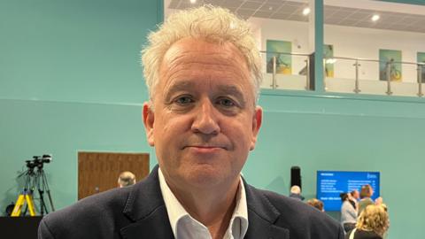Paul Dundas is wearing a suit on election night. He has grey hair and is standing in a sports hall.