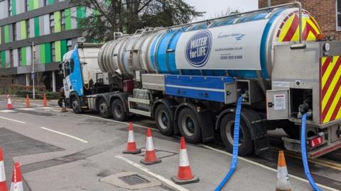 Water tanker outside green and gren building with blue hoses coming from it onto the road which has red an white cones along it.