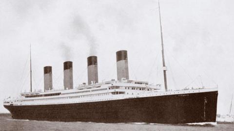 A black and white photo of RMS Titanic, a large ocean liner with four funnels, a black hull and a white upper deck. There are masts at the front and rear.