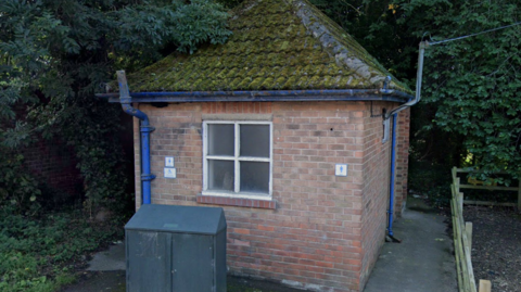 A small public toilet building with a pitched tiled roof and single square window made up of four panes. There is a sign for women and disabled people on the left and men on the right. Blue pipes run up both walls