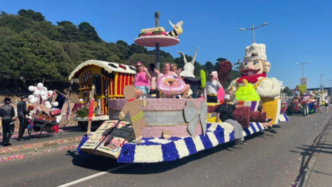 People stood on a colourful float which has a handmade cardboard gingerbread person as well as someone holding a large fake pink donut.

