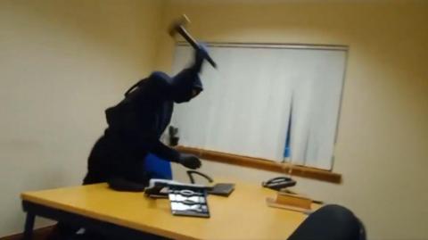 A person wearing black including a balaclava lifts a hammer before hitting a desk. There is a window with blinds drawn in the background.