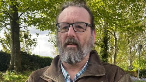 Jason Barker, a bearded man wearing glasses, standing in front of trees