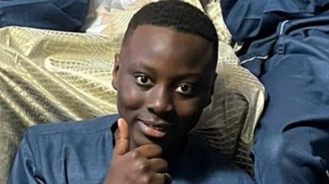 A young teenager sits smiling and giving a thumbs up to the camera. 