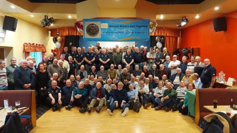 Ninety four members of the group at the annual get together. Some can be seen sitting on the floor of the venue with others standing behind. 