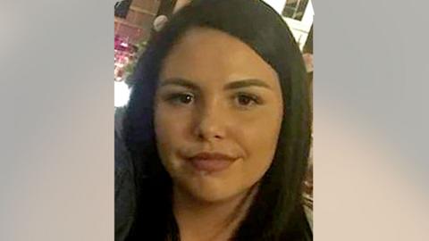 Alice Webb is smiling at the camera, she has dark hair and a piercing in her lower lip. She stands in front of an illuminated bar.