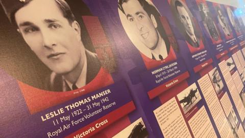 A close-up of a museum display showing black-and-white photos of RAF personnel who won the Victoria Cross and other honours. Each photo is mounted on a purple and red board, with text detailing their name, dates of birth and death, the medal they received and a description of their life and service. The closest photo shows the head and shoulders of a young man wearing an RAF uniform. The text reads: "Leslie Thomas Manser, 11 May 1922 - 31 May 1942, Royal Air Force Volunteer Reserve, Victoria Cross."