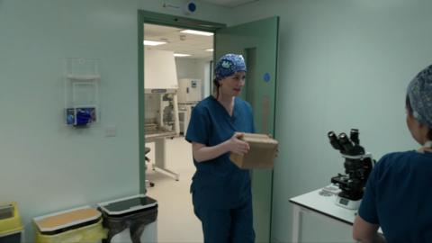 A woman in blue medical scrubs is seen carrying a plain box. She also is wearing a blue medical hair covering. She is in a room featuring a microscope, three bins typically found in a hospital, a light green door and turquoise walls. 