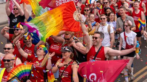A large group of Pride parade participants wearing red shirts march along a city street, waving rainbow flags and banners as colourful confetti falls around them.