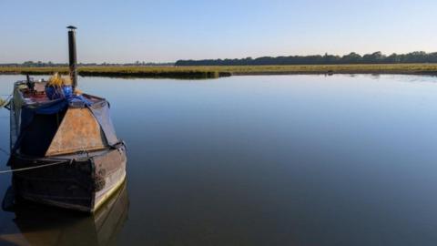 MONDAY - A body of water that is so still it doesn't have a single ripple stretches across the picture. In the water in the left of the frame is a rusting narrowboat with a chimney and a blue plant pot on top. On the far side of the water is a green meadow and on the horizon under the blue skies are a row of trees with the roofs of buildings behind.