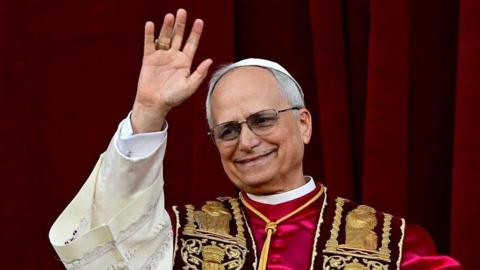 A photograph of Pope Leo waving to crowds. He is wearing an embroidered gown with red, cream and gold accents. 