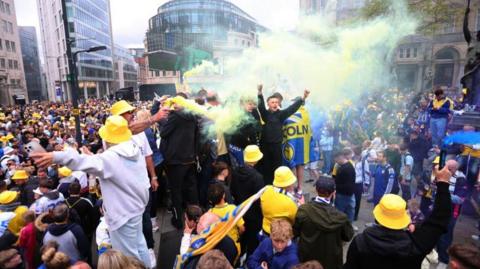 Lots of Leeds fans celebrating in kit, hats, with flares etc