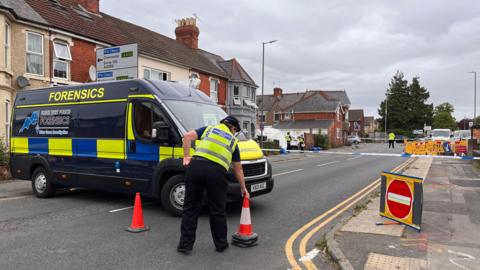 A forensics van with yellow and blue stripes on it. Forensics is written in yellow capital letters at the top. A police cordon can be seen on a residential street, consisting of a red no entry sign and white and blue police tape. An officer is standing next to the van, moving an orange traffic cone. The officer is wearing a yellow high vis jacket with police written on the back.