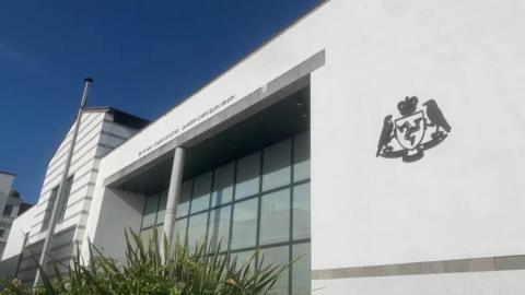 The exterior of the court building, a grand white structure, with large windows on a clear blue day, you can see the government crest of two birds facing the Manx flag.