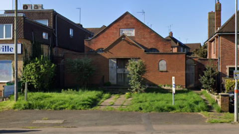 A red bricked building on a street between a home and a business. It has a pointed roof and looks like a chapel. The grass at the front is overgrown and the windows and doors are boarded up. 
