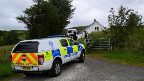 A police truck is parked at the entrance to the property. A police officer stands next to a metal farm gate and another vehicle and officer is parked near the house.