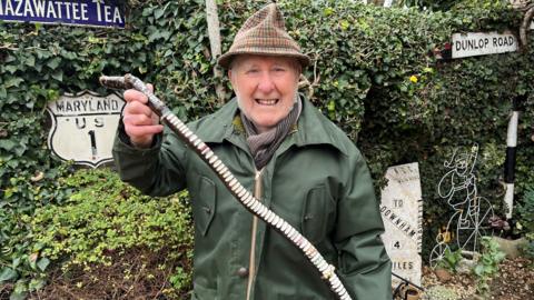 Colin Bedford, 88, wearing a green wax jacket overcoat, with a brown scarf underneath he has a tartan fedora hat and long grey whiskery sideburns. He is standing in his back garden with various old road signs behind him. He is holding a piece of tree branch about 1m (3ft) long with white notches cut into it, which represent every year he has done the walk.