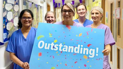 Five medical staff are dressed in scrubs and holding a blue banner saying the word outstanding! in a hospital corridor 