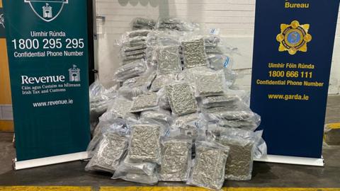 Cannabis is lying on the ground in a huge pile. The drugs are sealed in plastic bags with a blue zip lock. There are banners flanked at each side of the pile with contact numbers for the Irish tax and customs and also Gardi.