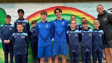 Two teenage boys in a light blue kit stand in the middle of a group of younger children in dark blue football kits standing against a wall with a rainbow painted on it.