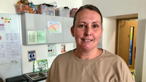 Kate Shivers-Poole wearing a brown sweatshirt with a sink and cupboards behind her, with posters and children's drawings stuck on the cupboards and baskets full of an assortment of items stacked on top of the cupboards