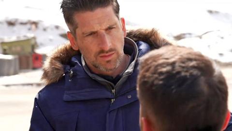 BBC correspondent Dan Johnson, wearing navy blue jacket, talking to a man with a blur hiding his face.