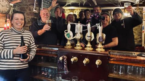 Emma Lomas holding a bottle behind the bar of The Crown Inn, with customers in the background raising pints