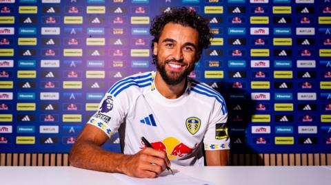 An image of Dominic Calvert-Lewin signing a contract while dressed in a Leeds shirt