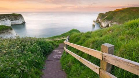 A steep pathway leads down to the sea with cliffs each side and an orange hue in the sky. The black path has a wooden fence alongside it and greenery is located each side of it.