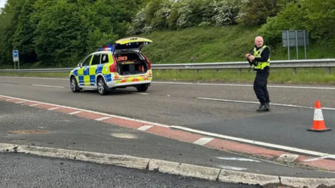 A police officer stands in the road on a section of the A1 between Morpeth and Berwick. A police car with an LED sign reading 'Road Closed' is parked in the road.