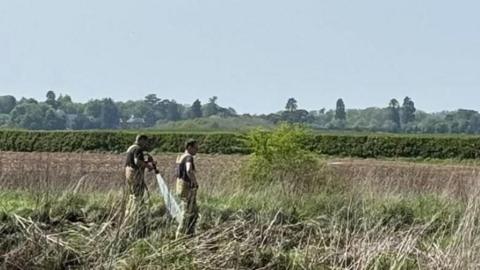 Two men in beige overalls standing in tall grass with a hedgerow in the distance