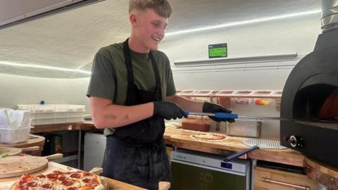 A teenage boy with dirty blonde hair is wearing a khaki green t-shirt, black gloves and a black apron. He is holding a utensil that is getting a pizza out of a wood-fired pizza oven in a small trailer
