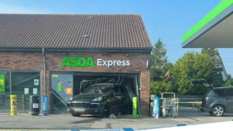 A Porsche Cayenne that is reversed into a shop window at an Asda Express petrol station. Police tape is across the forecourt. 